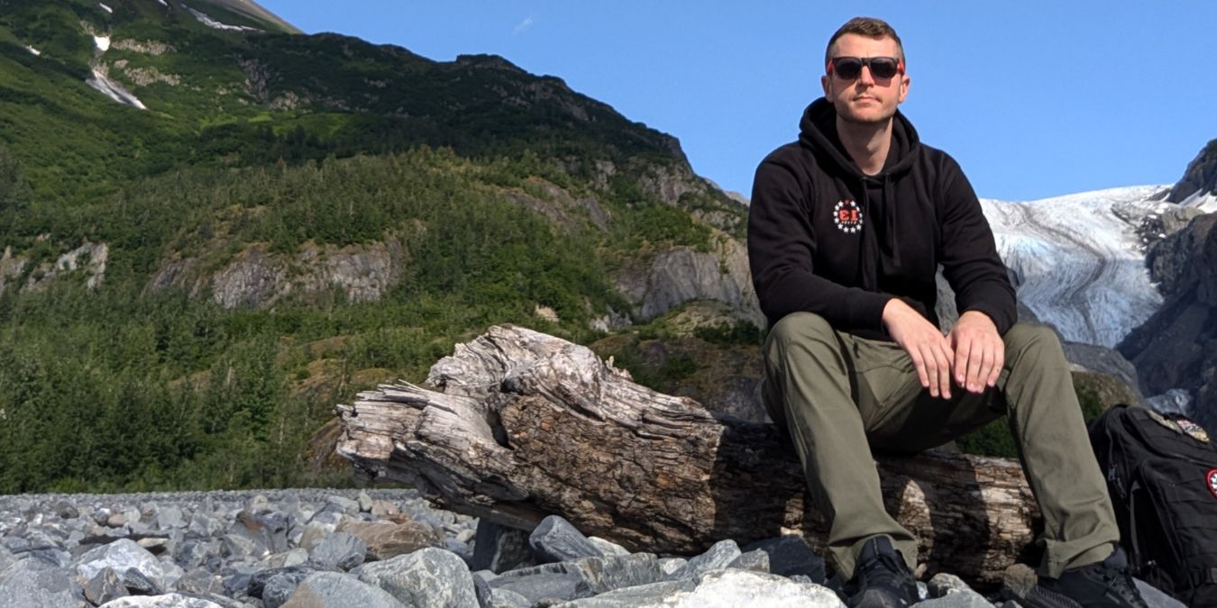 Sean from 13 Stars sitting on a log in Alaska with a 13 Stars Hoodie on