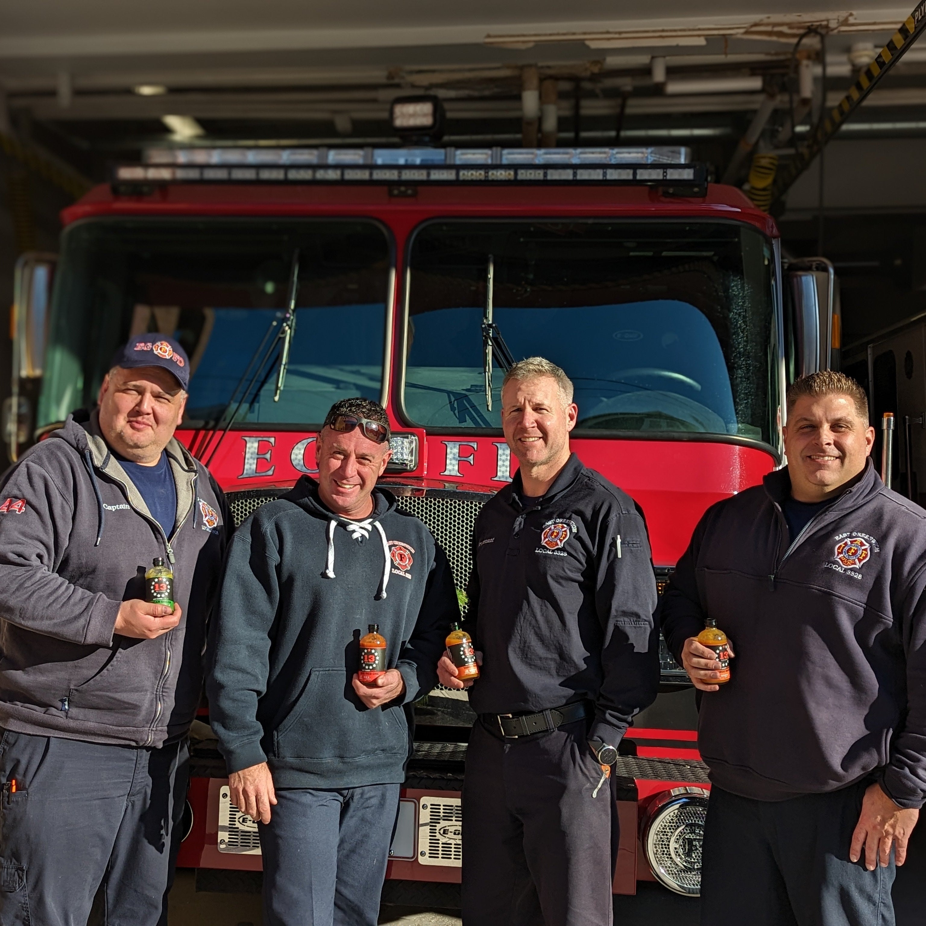 Four firefighters standing in front of a fire truck holding 13 Stars Hot sauce.