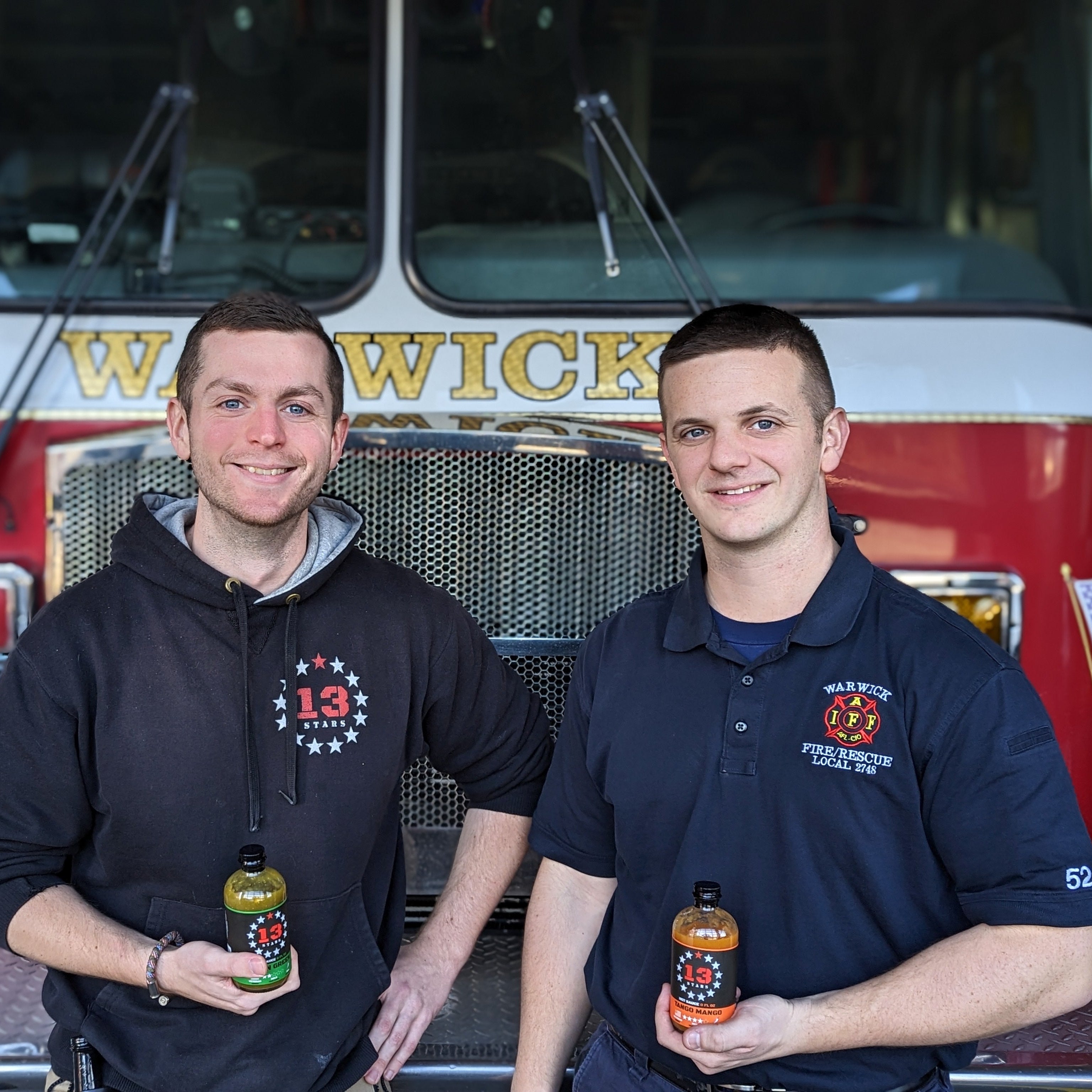 Two men standing in front of a fire truck holding bottles.