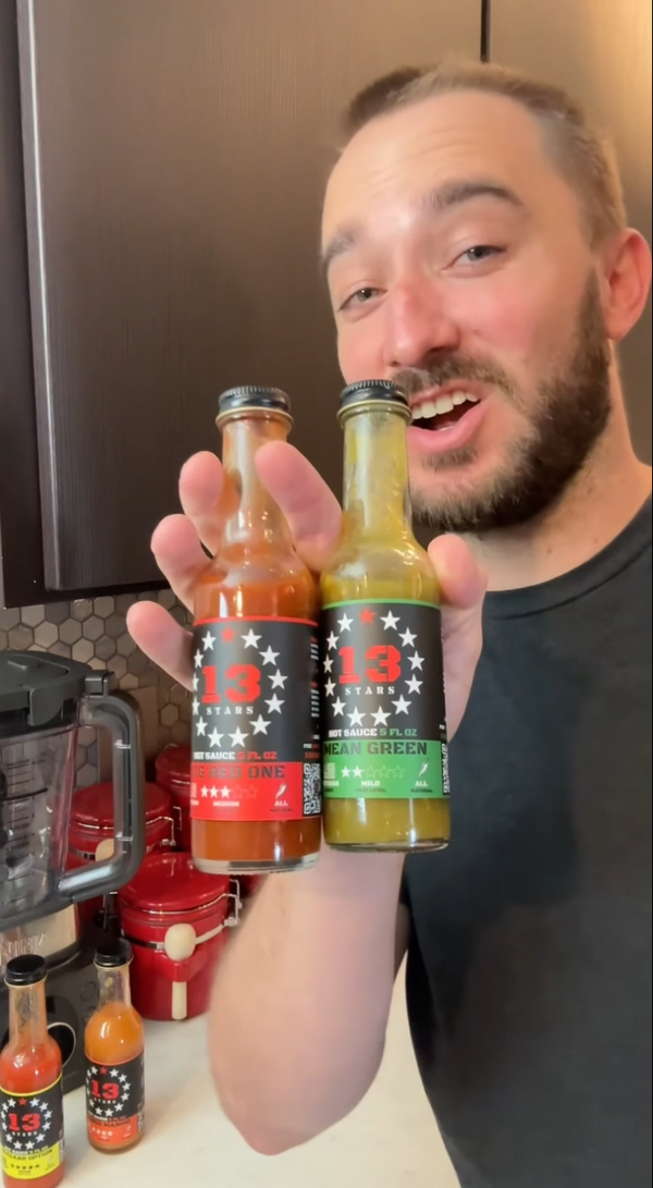 Man holding two bottles of '13 stars' brand hot sauce in a kitchen setting